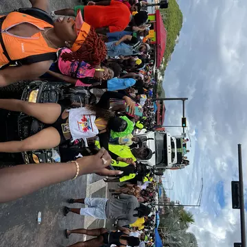A woman poses on a small motorized cart while a large group of people walk pass during J'Ouvert in St Thomas. There is a large truck in the background.