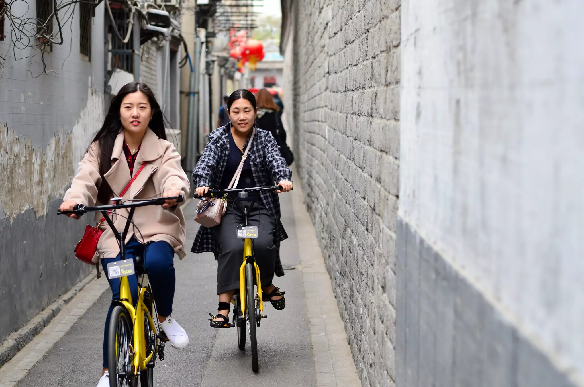 Two young woman ride yellow bikes in a narrow alley of a city.
