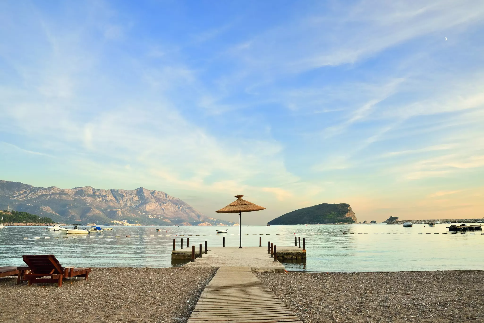 A small wooden jetty leads out from a pebbly beach into a cove with a large rocky islet in the center.