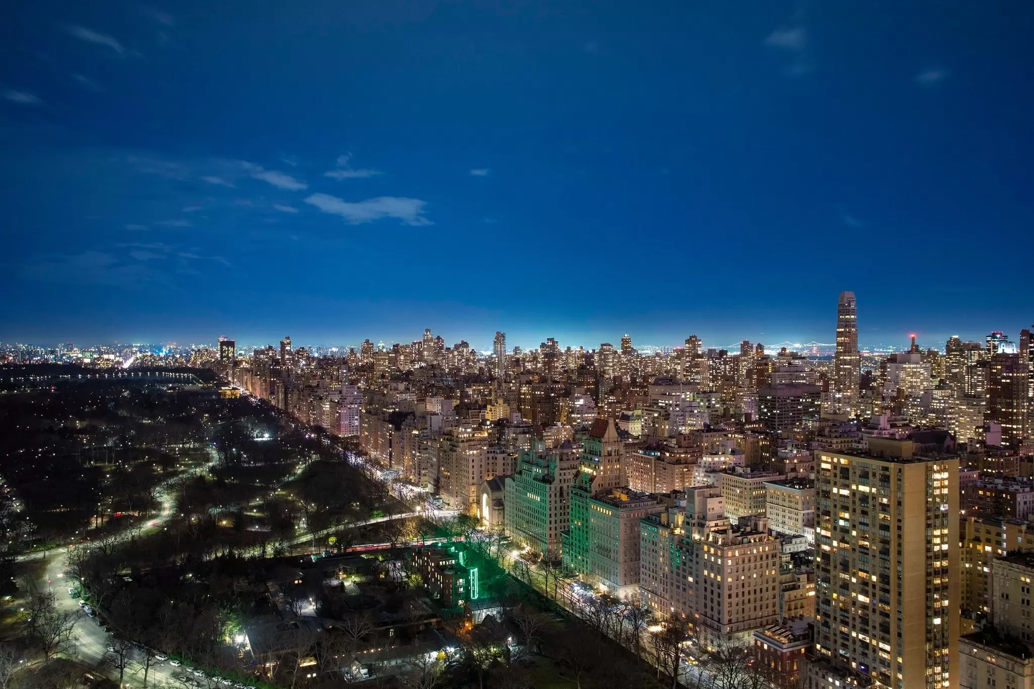 A dense city with buildings lit up and a darkened park at night from a rooftop perspective.