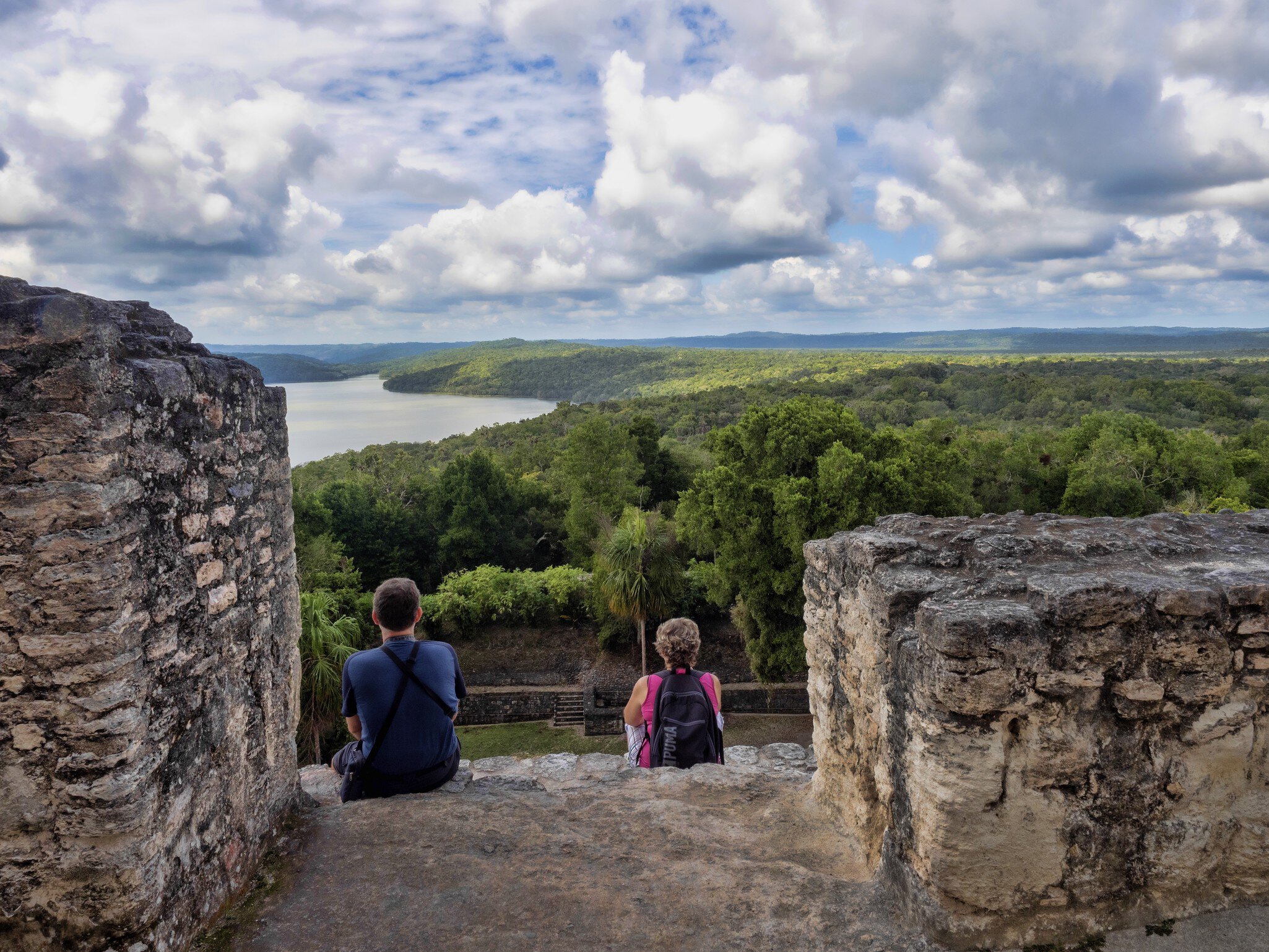 Two visitors seen from behind sit between weathered stone walls, looking out over jungle canopy to a lake under clouds