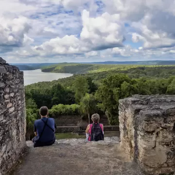 Two visitors seen from behind sit between weathered stone walls, looking out over jungle canopy to a lake under clouds