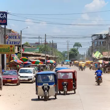 Red and blue three-wheeled mototaxis driving down a street past shop signs and striped umbrellas in Puerto Maldonado, Peru.