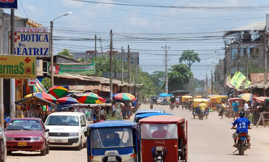 Red and blue three-wheeled mototaxis driving down a street past shop signs and striped umbrellas in Puerto Maldonado, Peru.