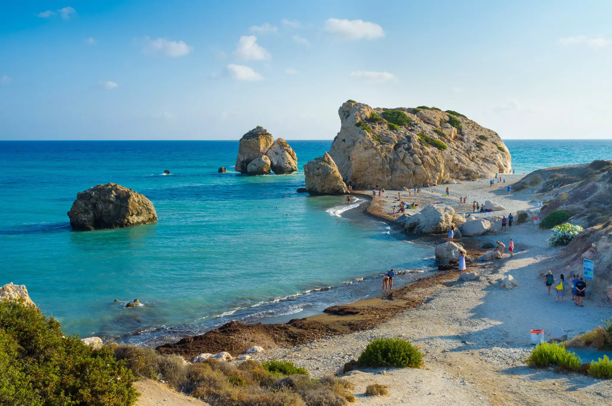Tourists mill around a stony clean beach admiring several huge rocky outcrops at sea