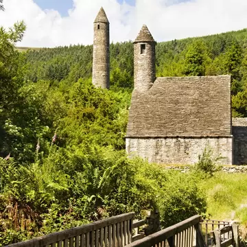 Tall stone round tower and a small stone church with a steep roof and belfry, seen past a wooden footbridge and ferns.