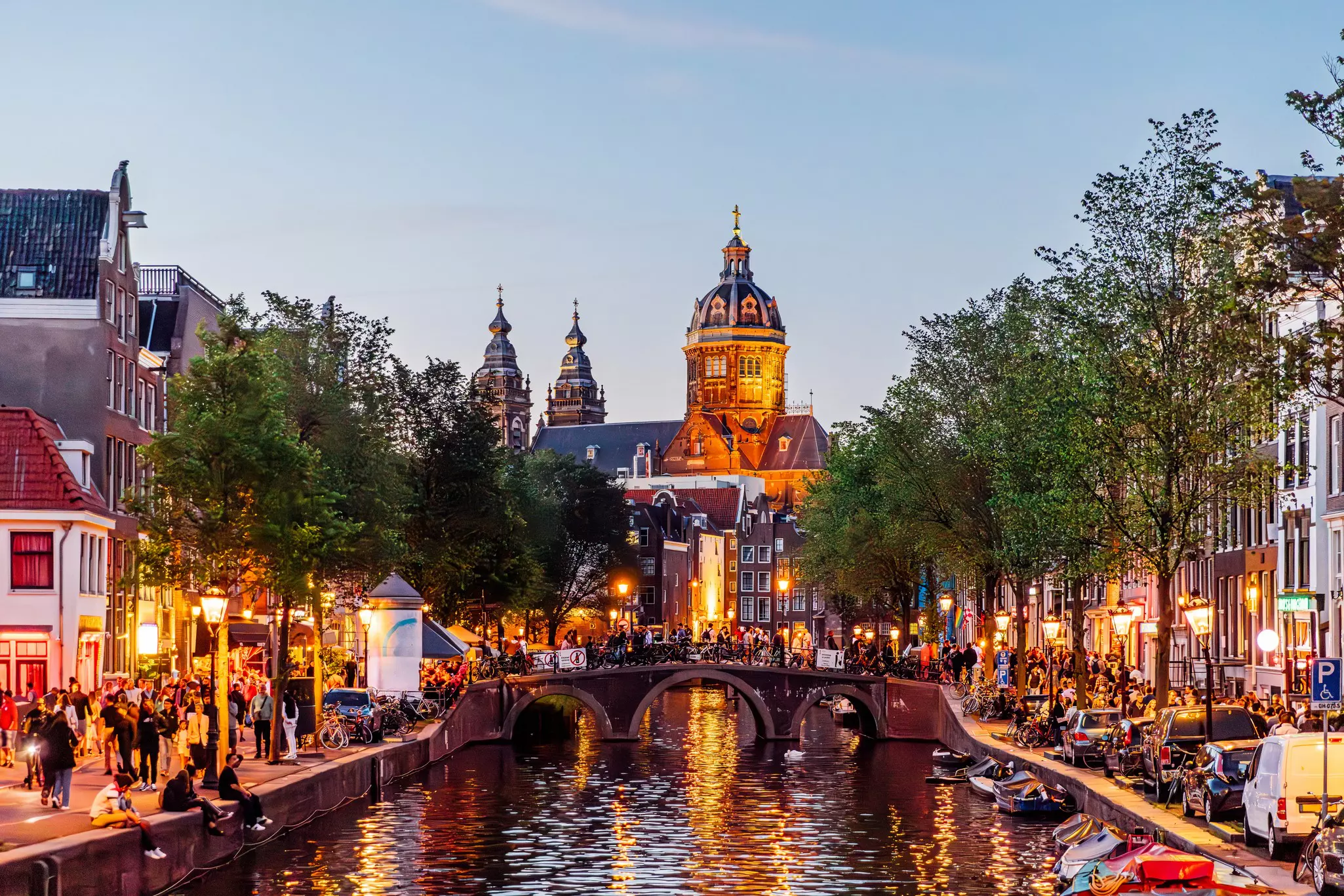 A busy neighborhood with people standing outside buildings either side of a canal at dusk
