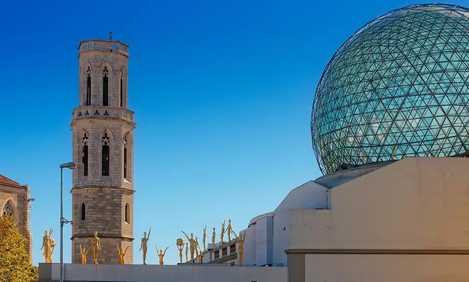 Figueres: Dalí Theatre-Museum's glass geodesic dome and gold figures on its roofline beside a Gothic stone bell tower