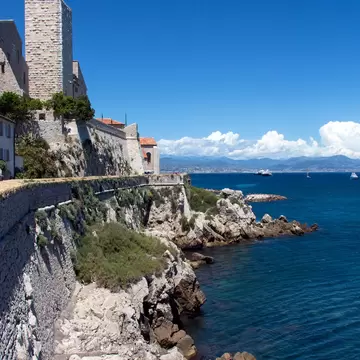 Stone ramparts and square castle tower of Antibes above rocky shore and deep blue sea, with mountains across the bay.