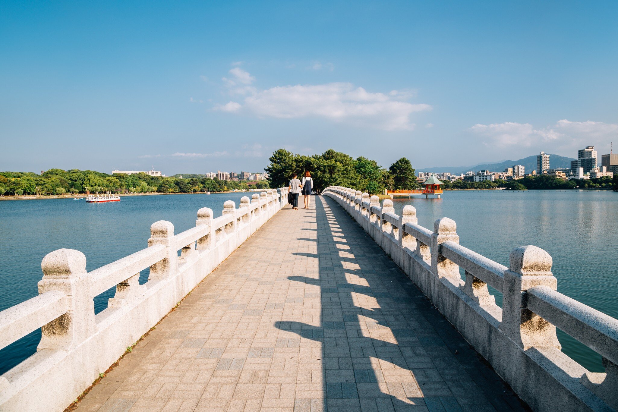 Couple walking a white stone bridge across the lake in Fukuoka's Ohori Park, with city towers beyond