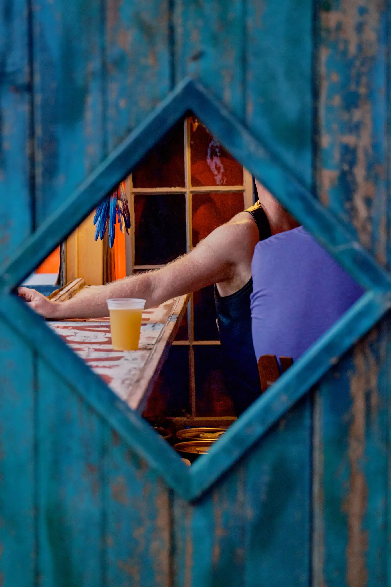 A man's arm and beer on a bar seen through a diamond shaped porthole in a blue door