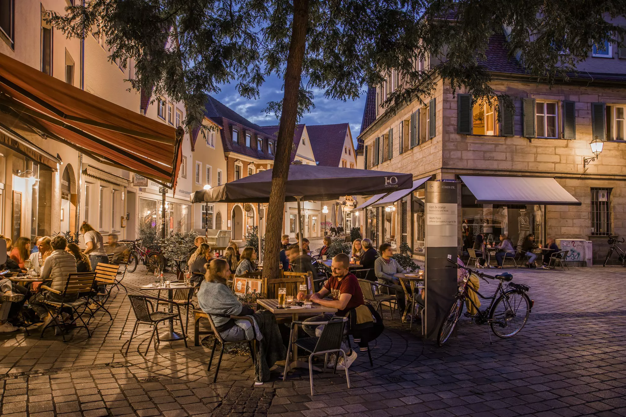 A nighttime scene shows many people sitting at outdoor tables at a cafe in Bayreuth, Germany, with a cobblestoned square and bicycles around.
