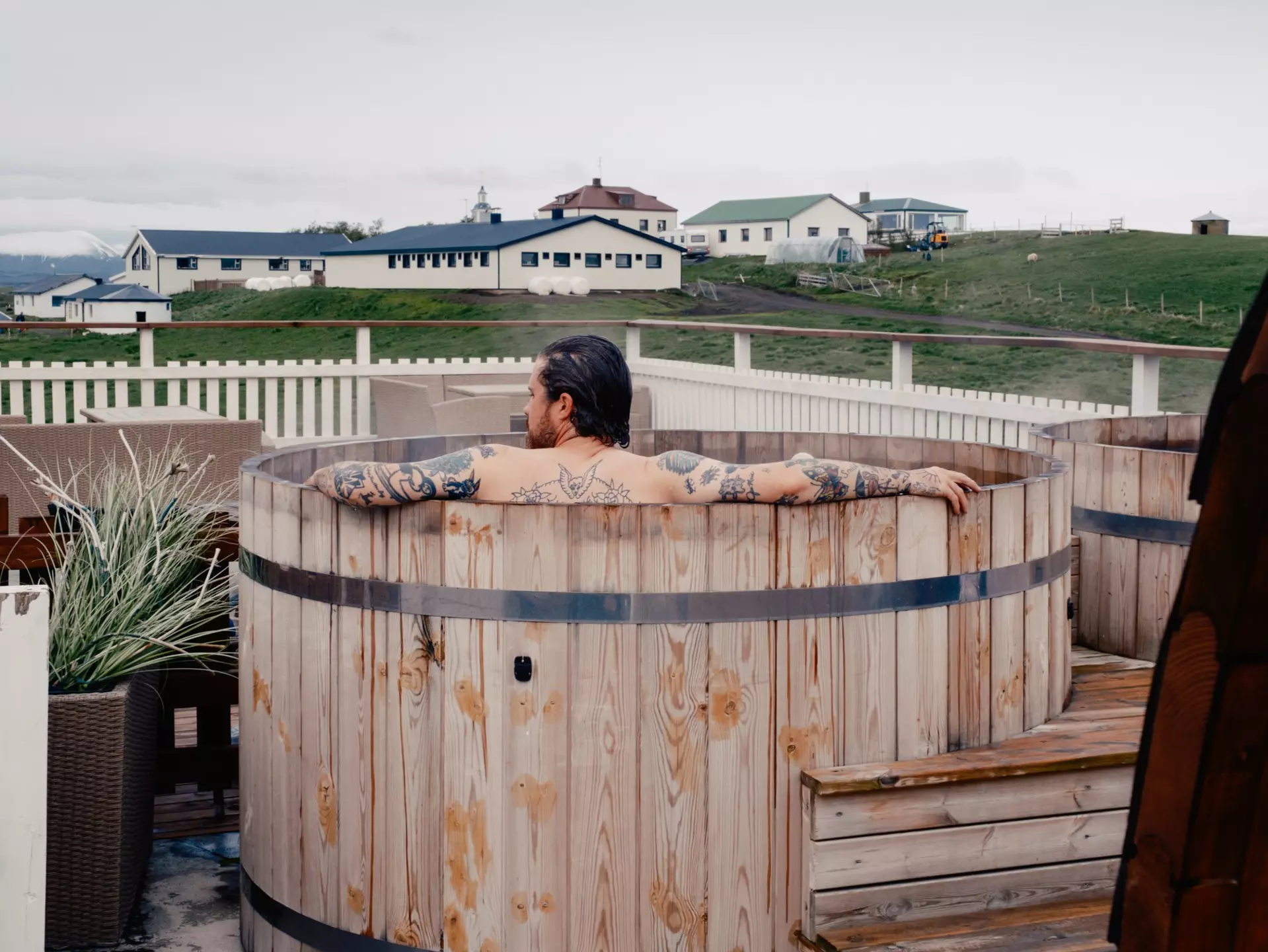 A person in a wooden hot tub; a large, low building is across a grassy area.
