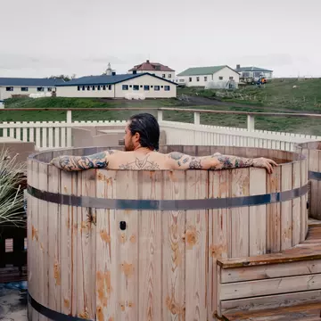 A person in a wooden hot tub; a large, low building is across a grassy area.