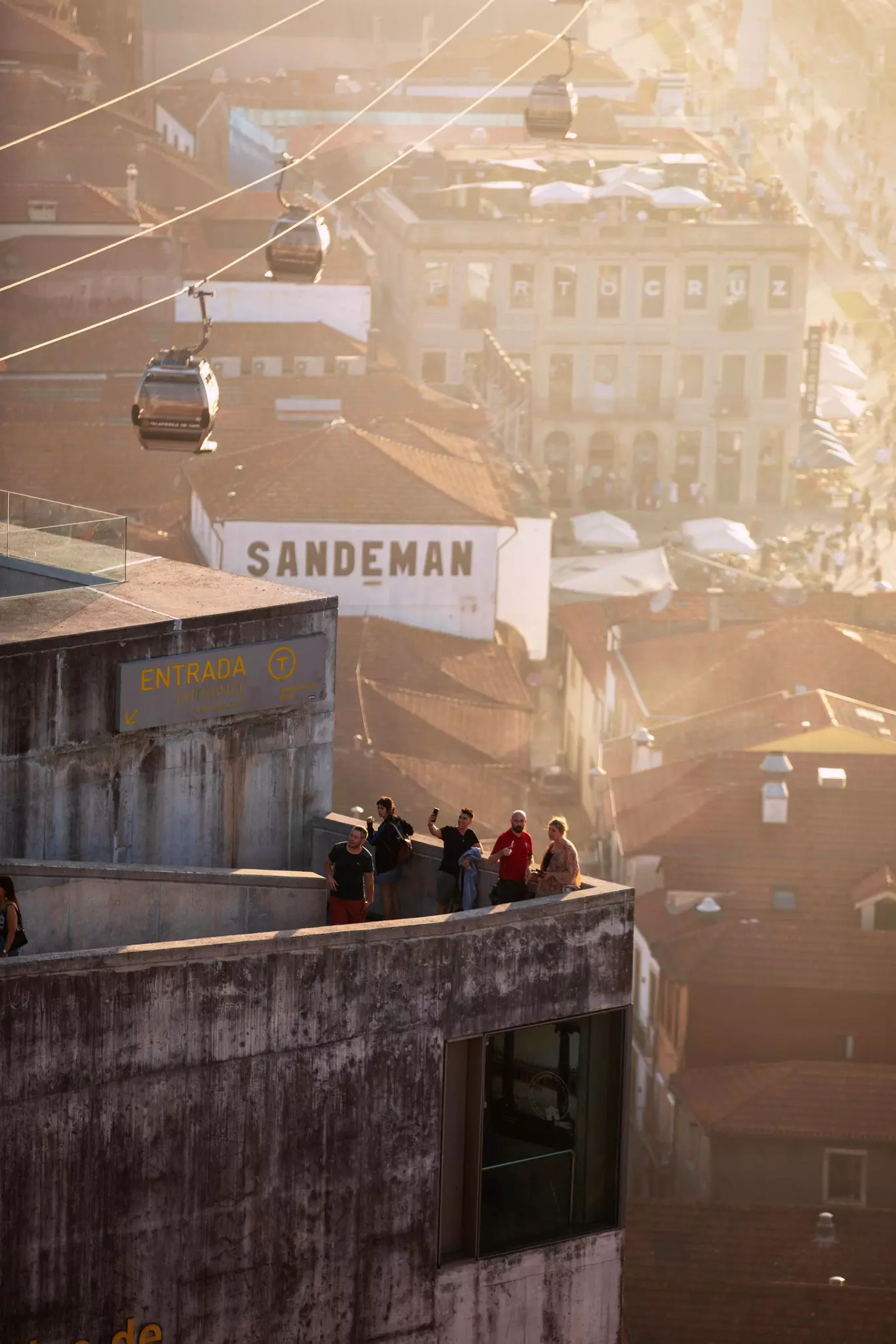 People at the edge of a railing at sunset, high above the buildings below; three cars on the cable of gondola are in the background. 