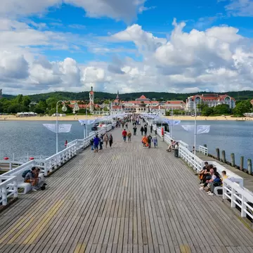 People walk and sit along a broad wooden pier with white railings leading to a beach and red-roofed buildings, Sopot, Poland.