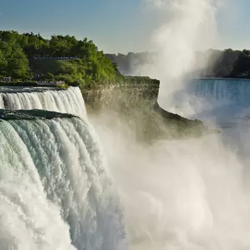 A broad waterfall pouring over a rock lip in white sheets, spray rising in a tall plume, with wooded clifftop viewing areas above.