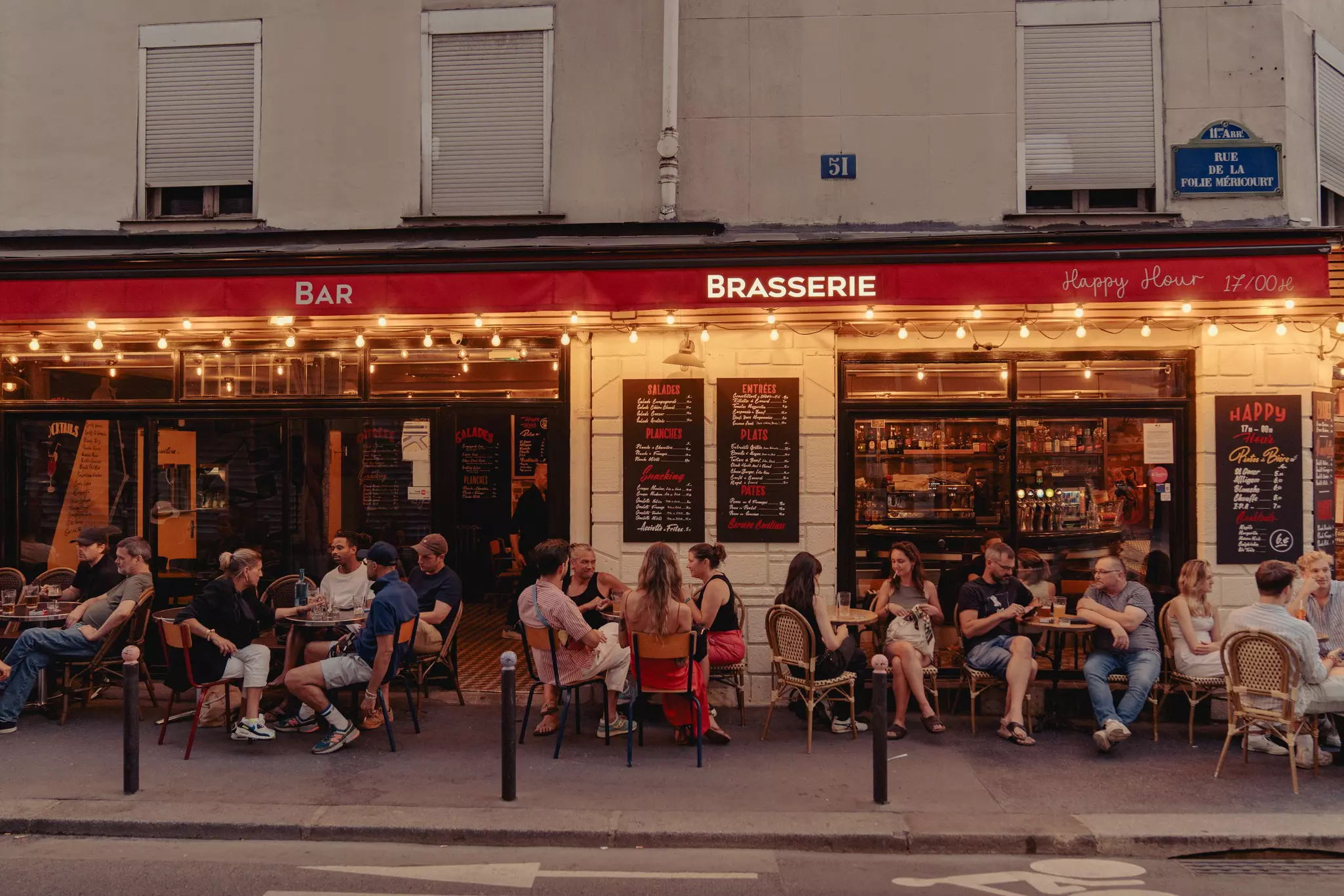 People sit at outdoor tables outside a brightly lit bar at dusk.