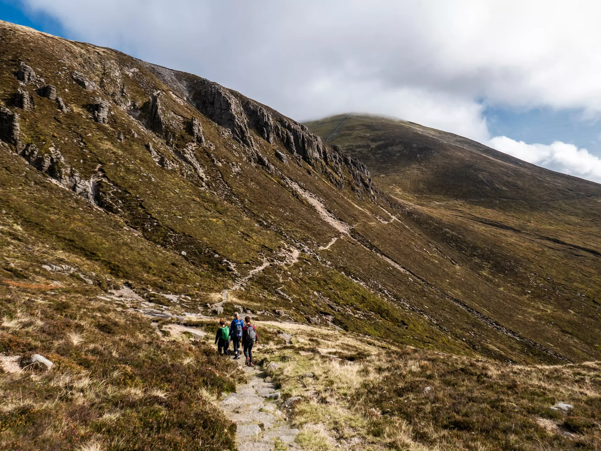 A wide shot of hikers following a trail along a ridge of mountains covered in shrubs.