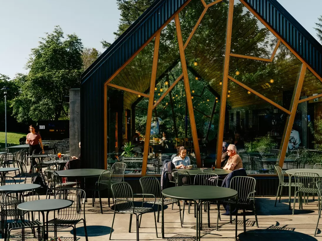 Black A-frame café with angular timber-framed glass walls, people at outdoor tables beside a flower bed in Lystigarðurinn