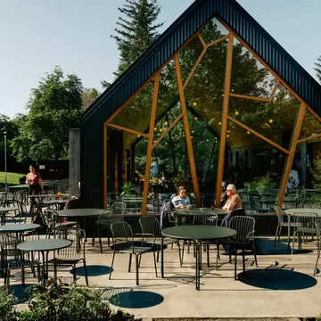 Black A-frame café with angular timber-framed glass walls, people at outdoor tables beside a flower bed in Lystigarðurinn