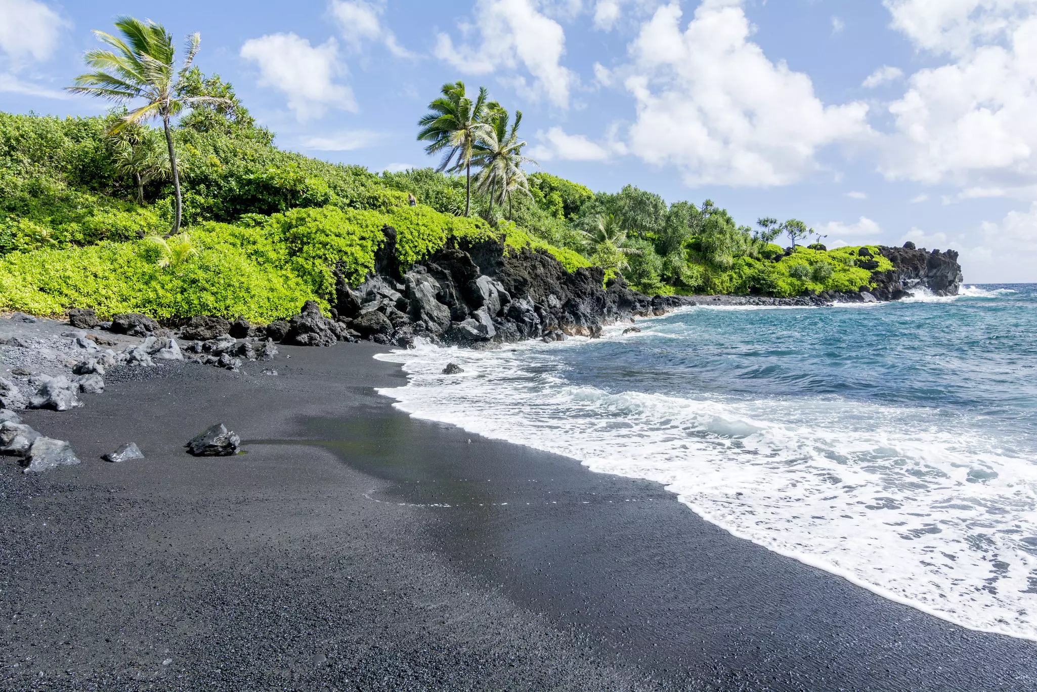 White foam edging blue water coming ashore on black sand in Maui, Hawaii, with a rocky shoreline that has very green trees and bushes