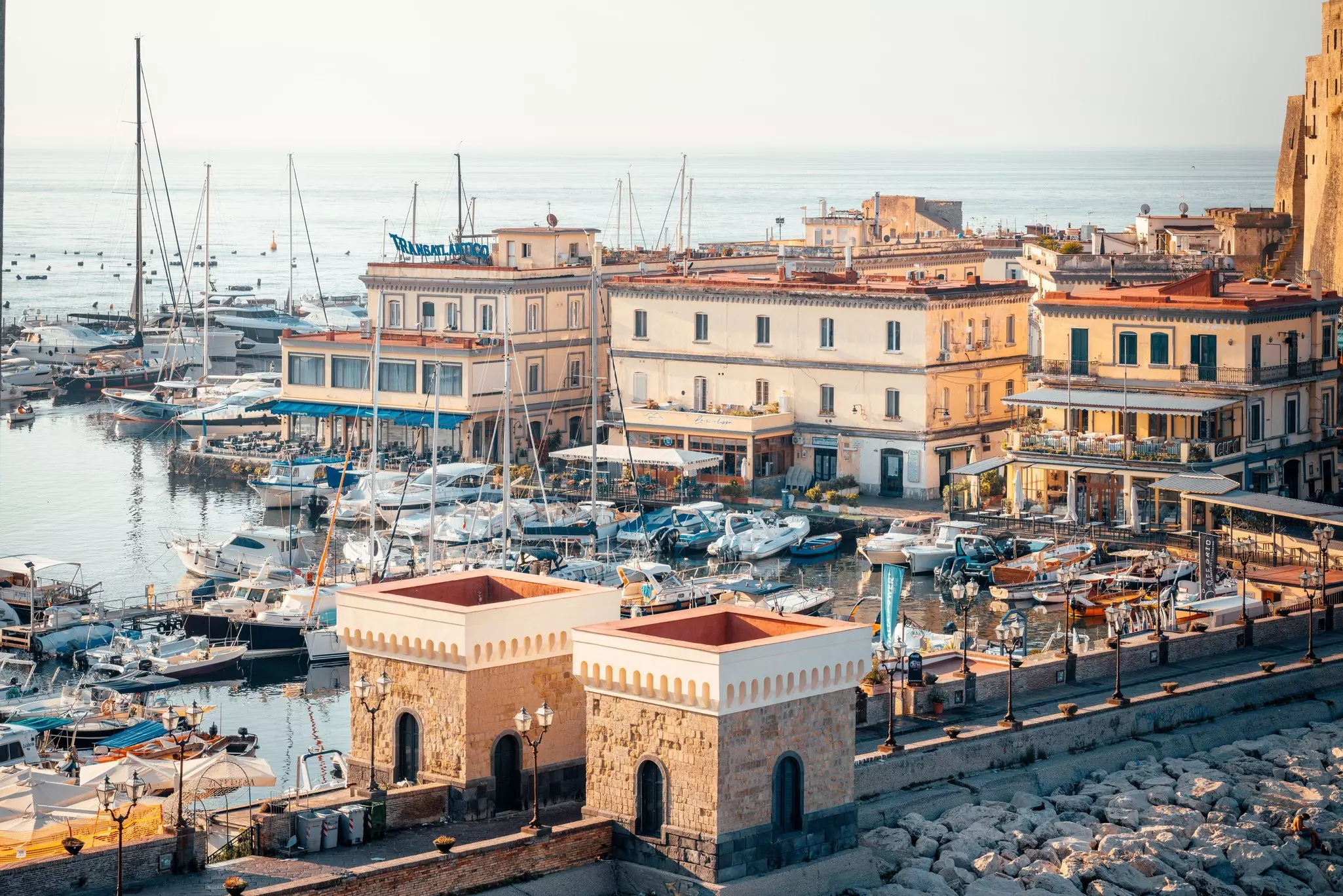 An aerial view of boats in a small harbor in Italy; there are low buildings on the shore.