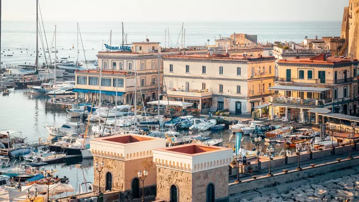 An aerial view of boats in a small harbor in Italy; there are low buildings on the shore.