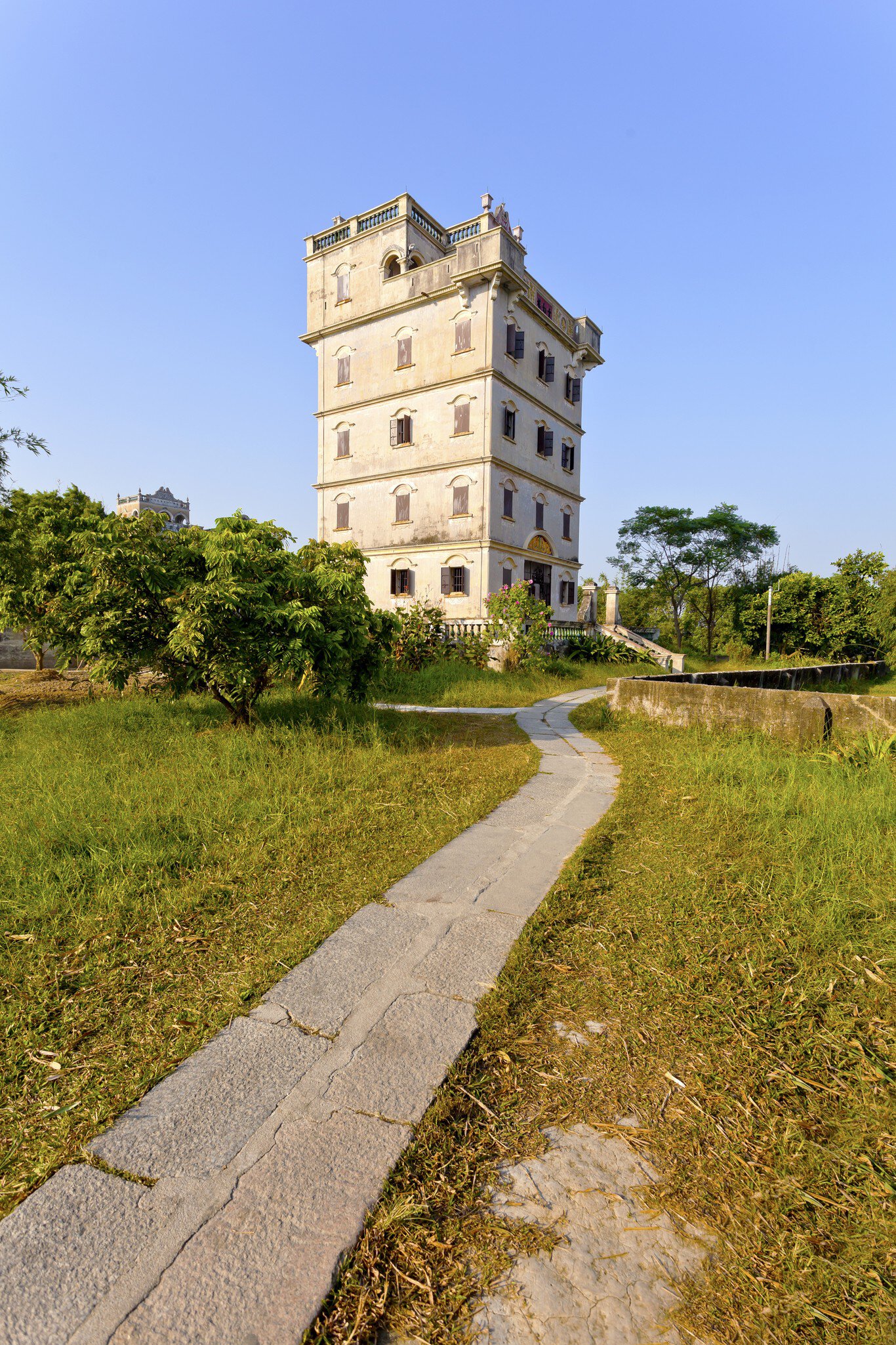Tall pale fortified tower house with shuttered windows and a balustraded top, a stone path leading to it, Kaiping.