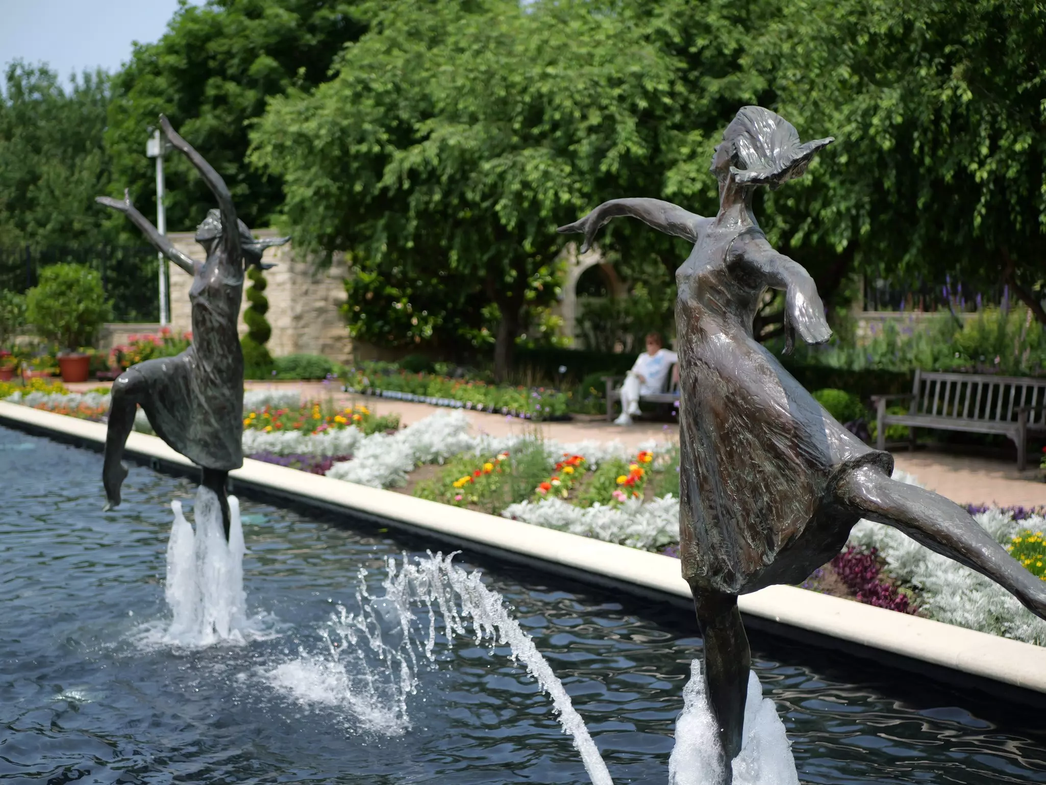 Sculptures of dancing women form part of a fountain in a park with trees and flower plantings.
