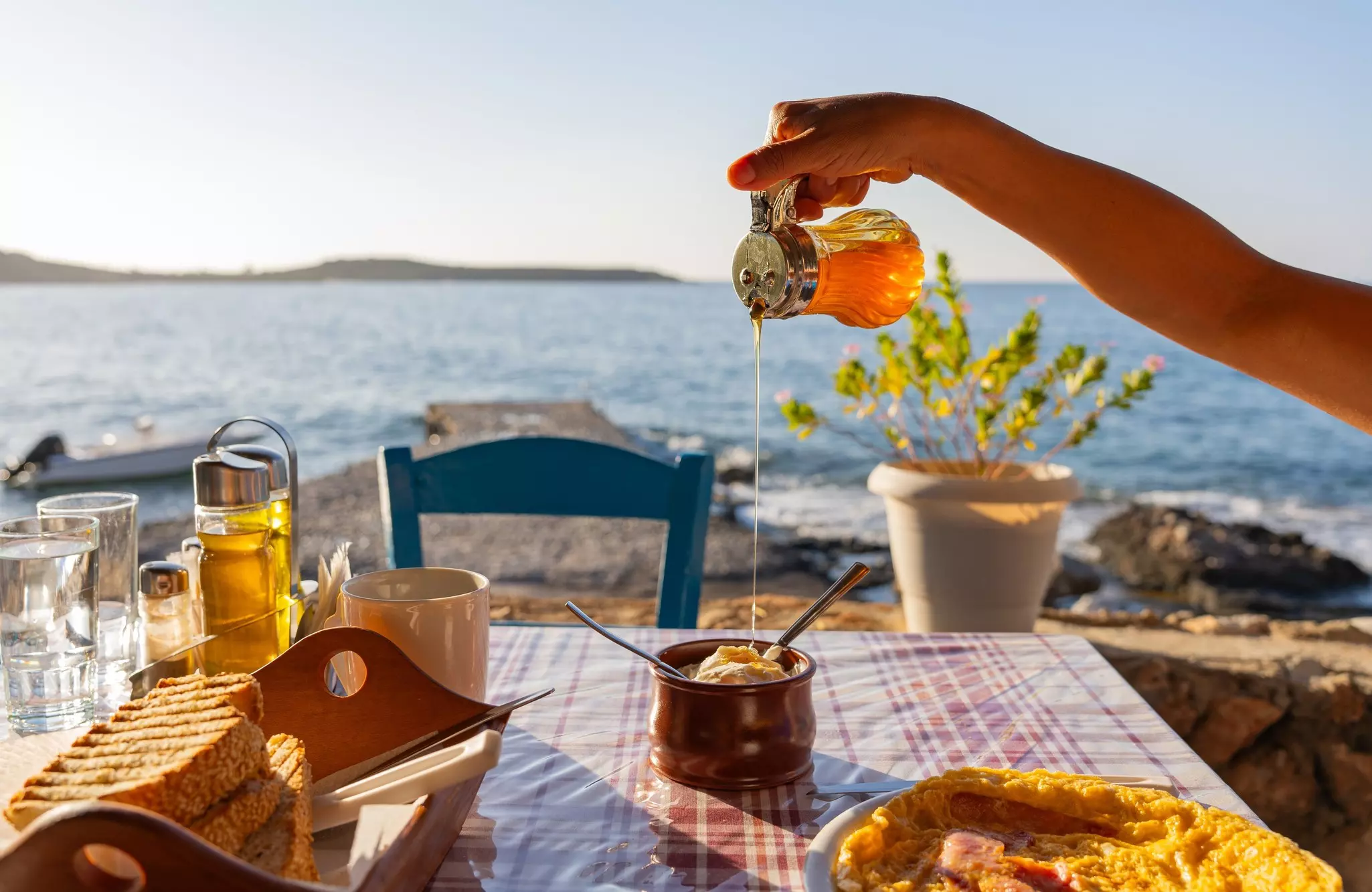 Greek tavern table at sunrise with a full breakfast and hand placing honey on yogurt and with the Aegean sea in the background.