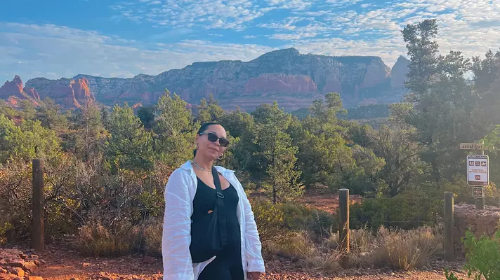 A person stands on a trail with trees and reddish rock formations in the background.