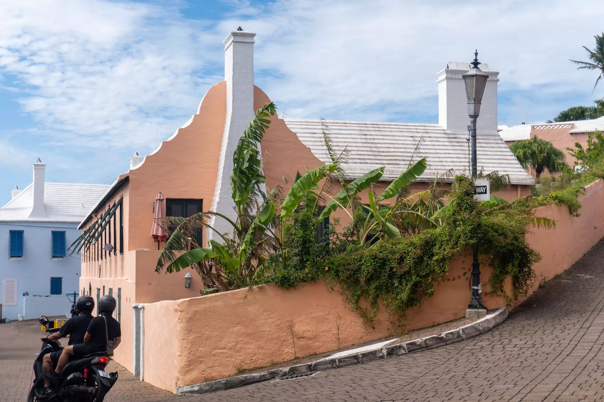 Two people on a moped drive on a cobblestone street past a peach-colored stucco house on a corner with a white house in the distance.