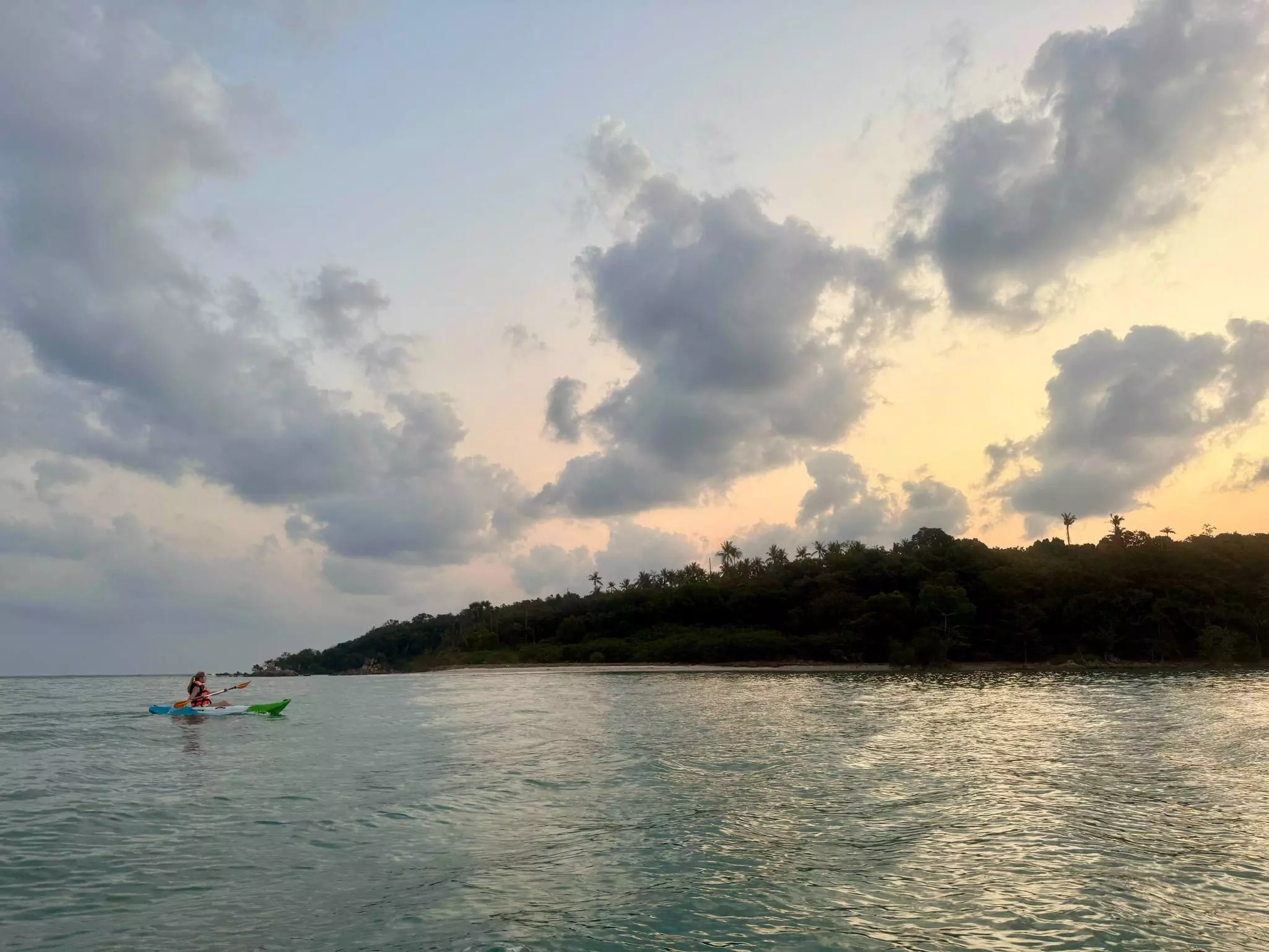 A kayaker paddles along near the coastline of an island as the sun starts to lighten the gray sky