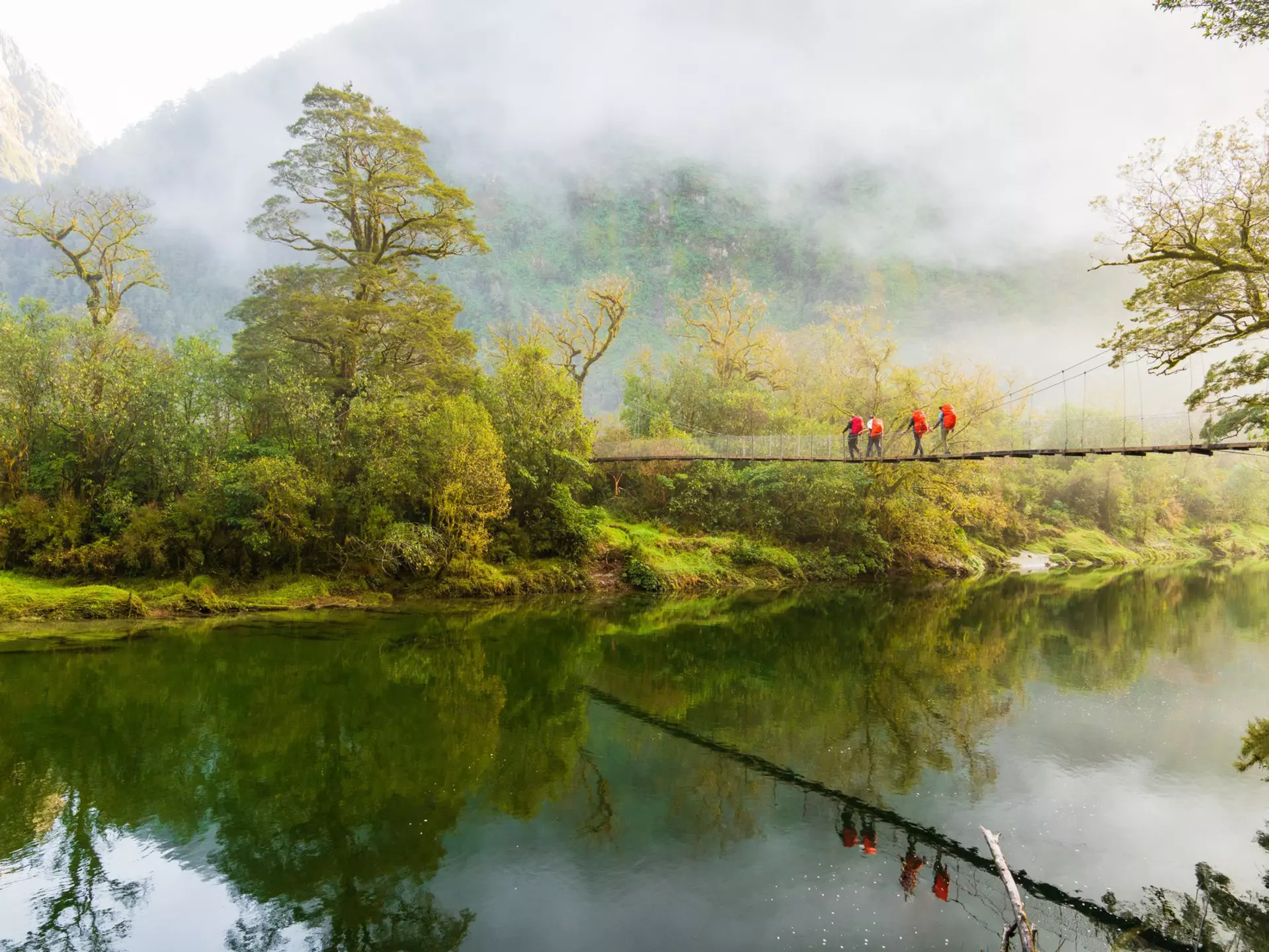 Hikers cross a bridge suspended above a river on a misty day