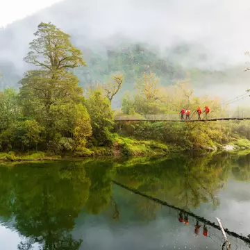 Hikers cross a bridge suspended above a river on a misty day