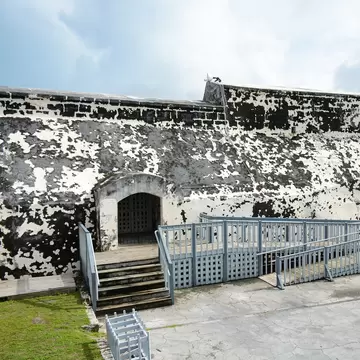 Mottled black-and-white stone fort walls with an arched gateway and wooden ramps in a paved courtyard, Nassau