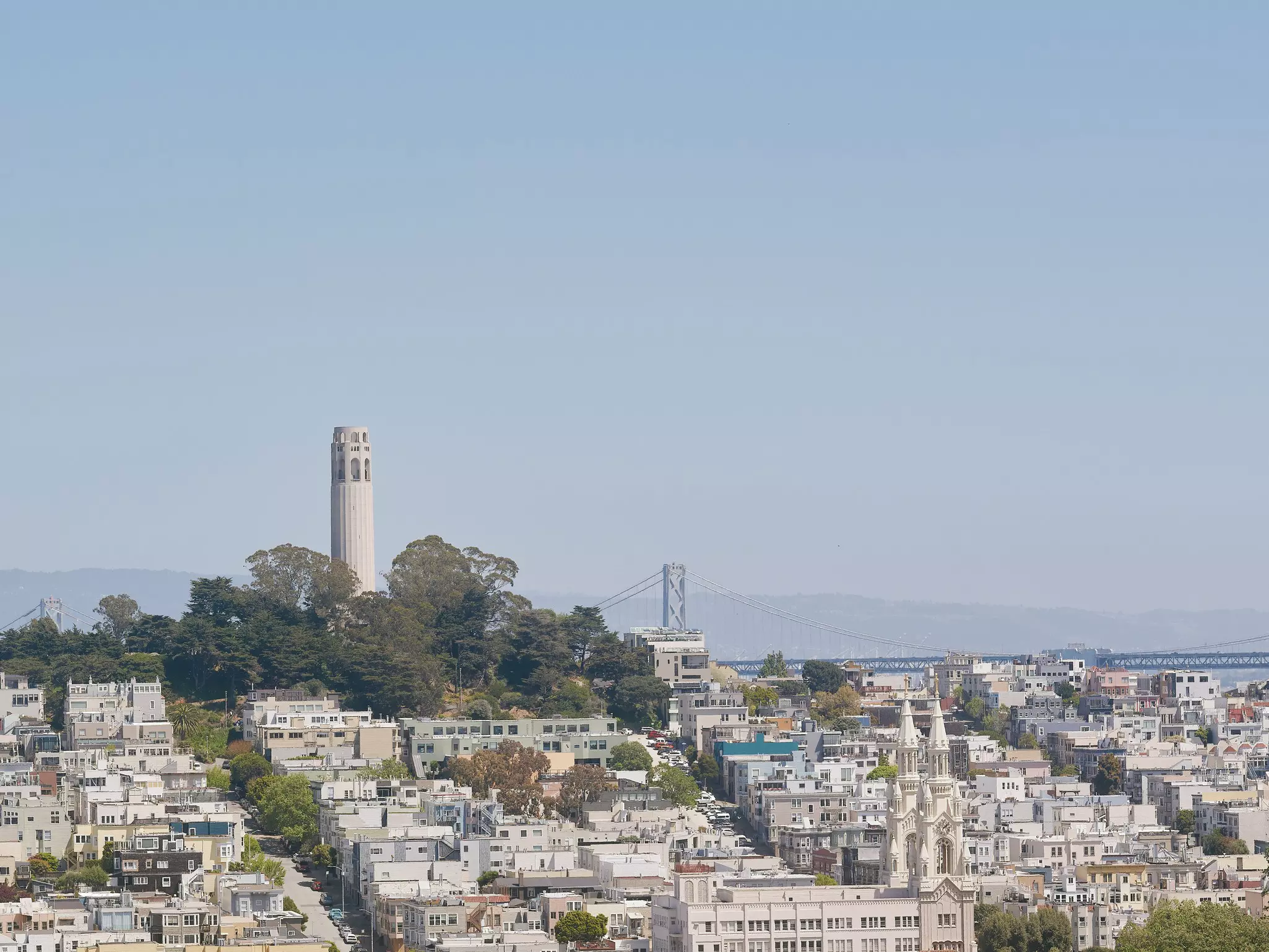 A tall tower surrounded by trees on a hill over neat city blocks full of square buildings a bridge in the distance