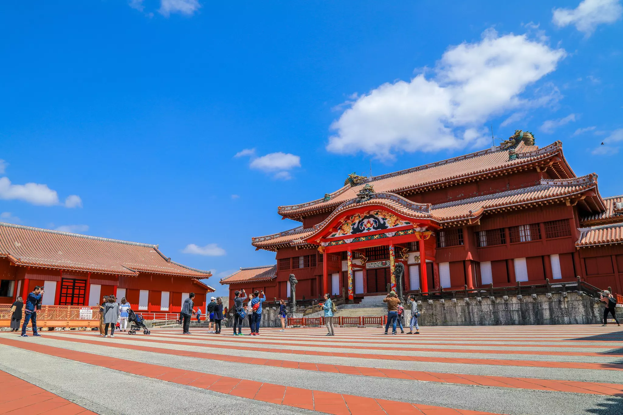 People mill around the courtyard of two large, red traditional Japanese buildings