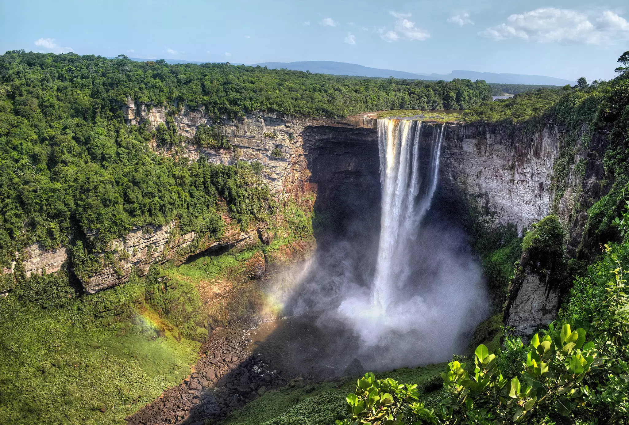 Aerial view of a large waterfall pouring into a basin from a rocky cliff on a sunny day.