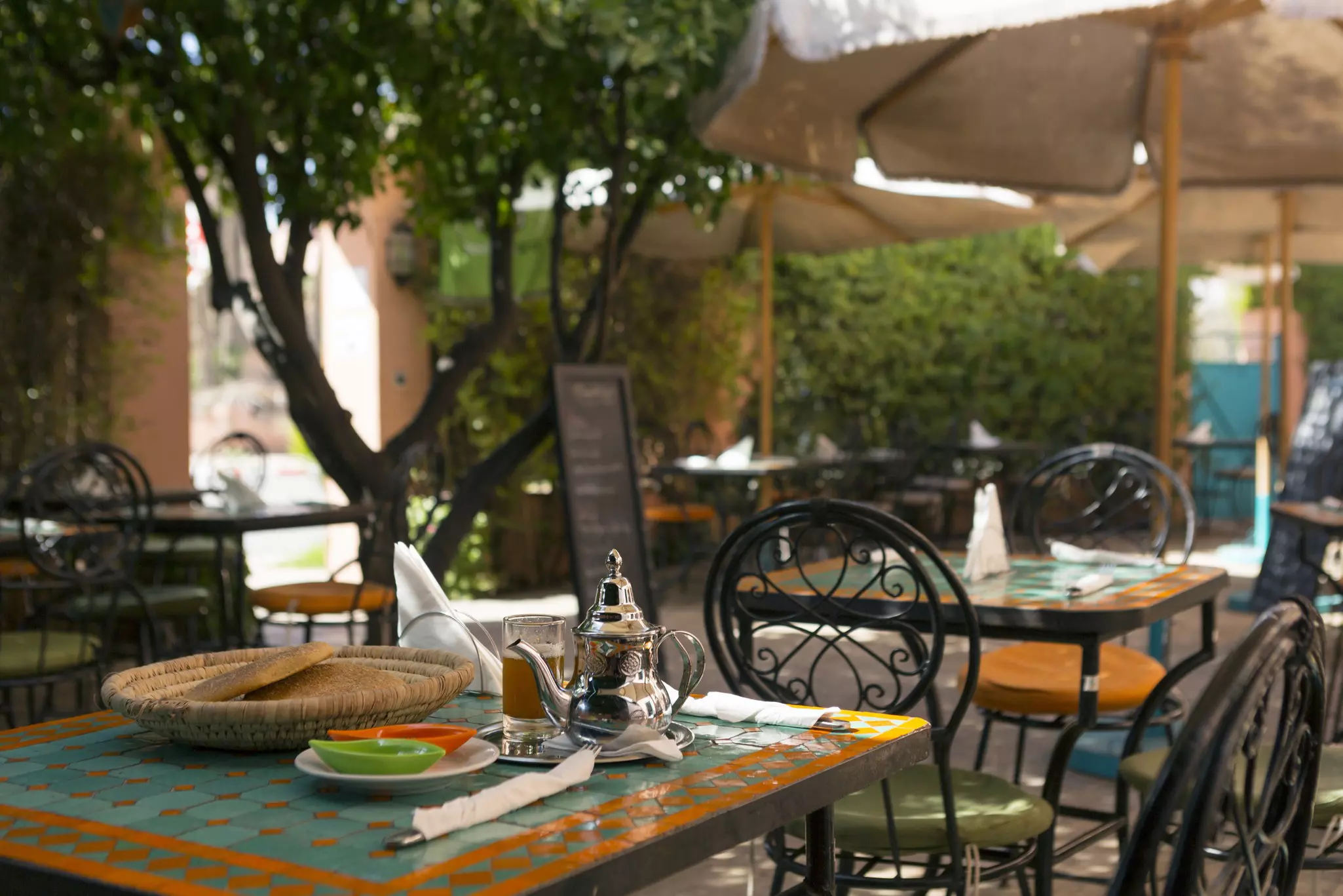 A table in a restaurant courtyard with a basket of flatbread and a silver pot next to a glass of tea.