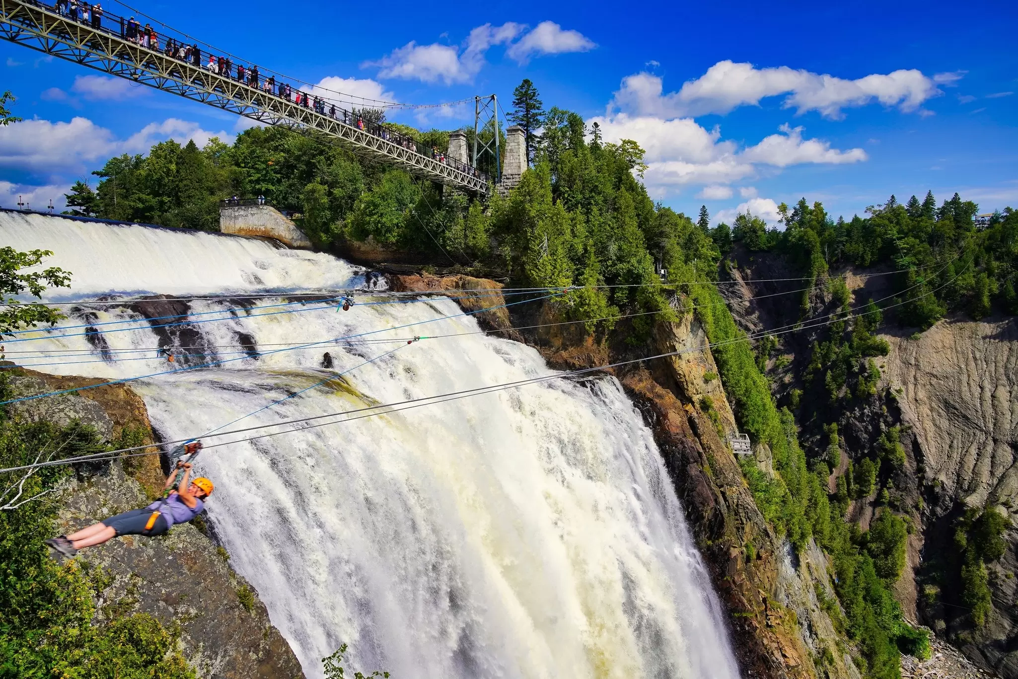 Huge, steep waterfall with green forest either side and a person ziplining across the front of it