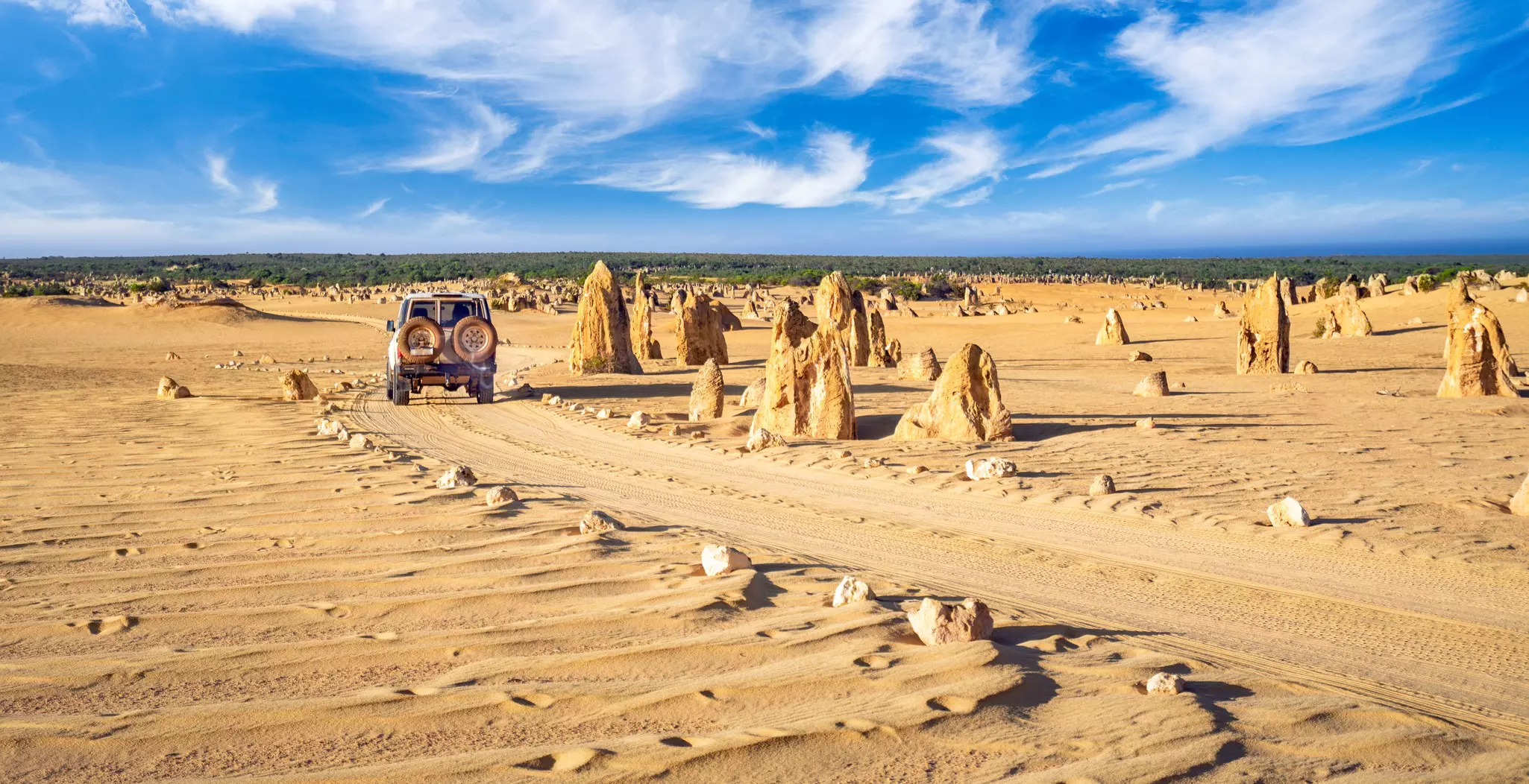 A four-wheel drive vehicle follows a sandy track through pointed rock formations that jut straight out from the sand.