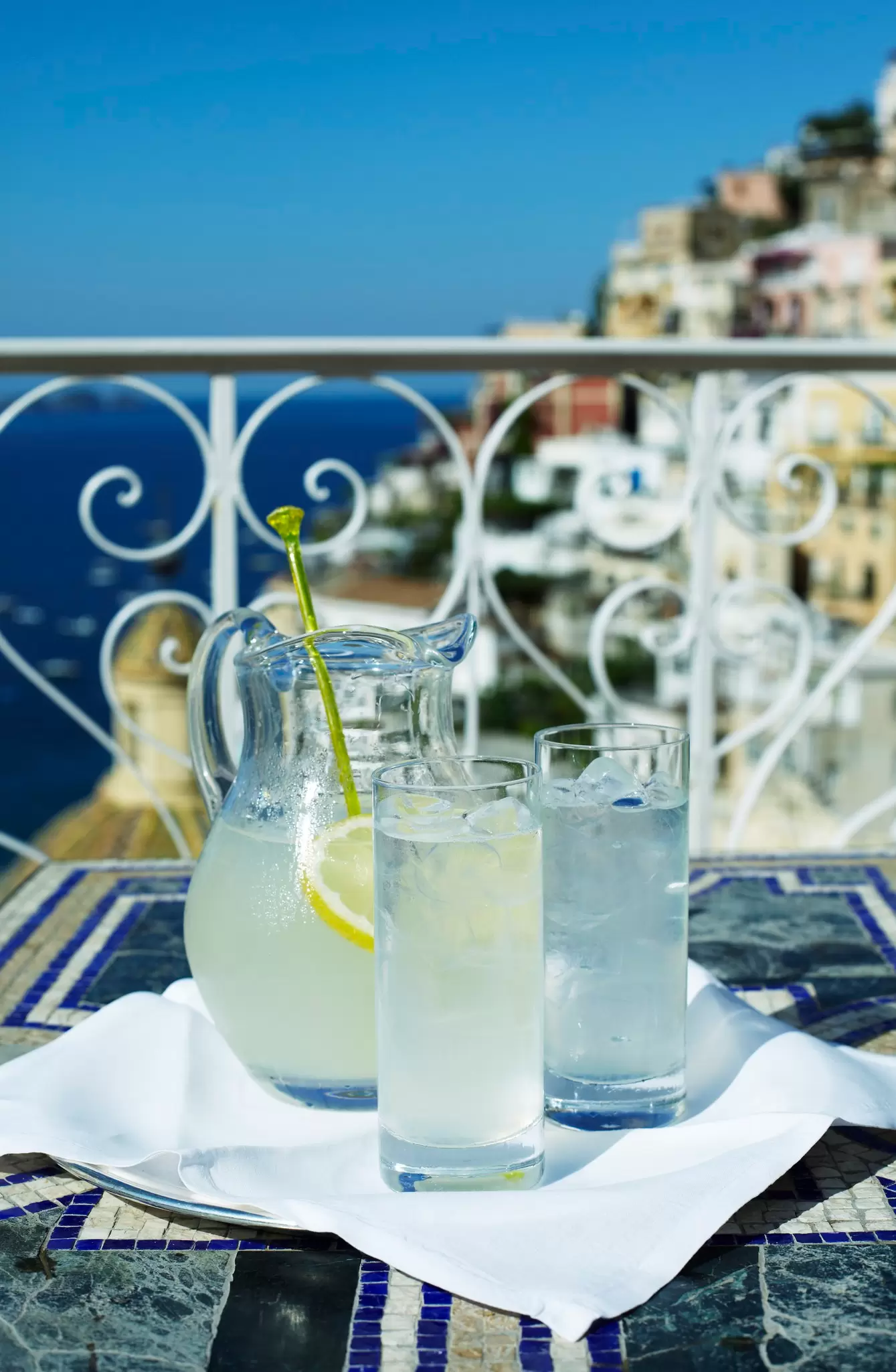 Glasses of lemonade on a balcony table with wrought-iron railing and Positano's hillside houses beyond.