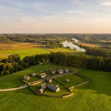 Aerial view of a palisaded wooden village on a grassy hillfort, with forests and the winding Neris River beyond at sunset