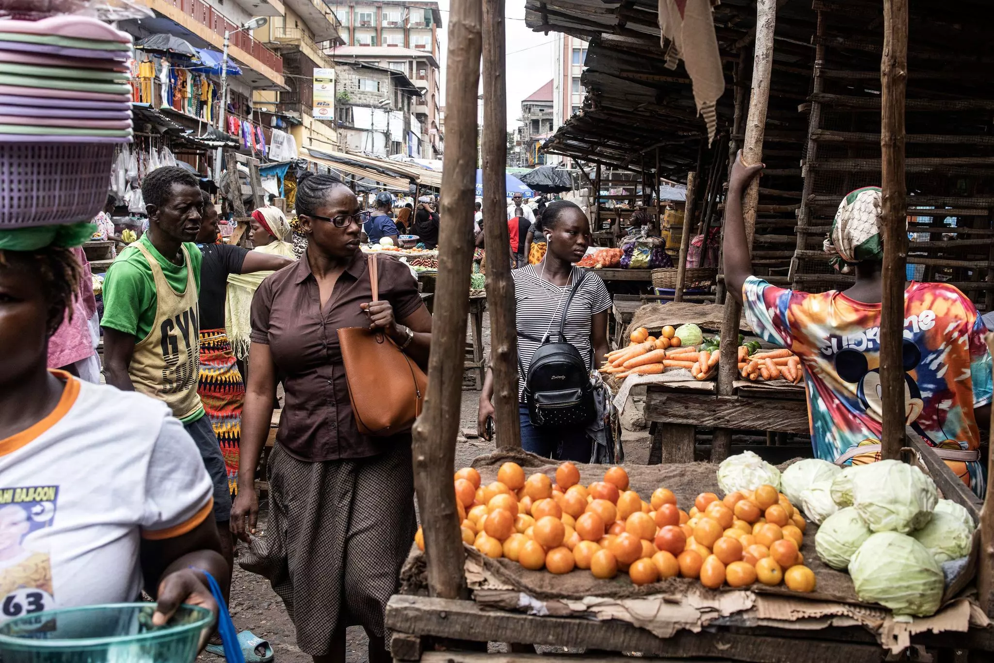 People look at fresh produce on display at an outdoor market in a crowded urban neighborhood.