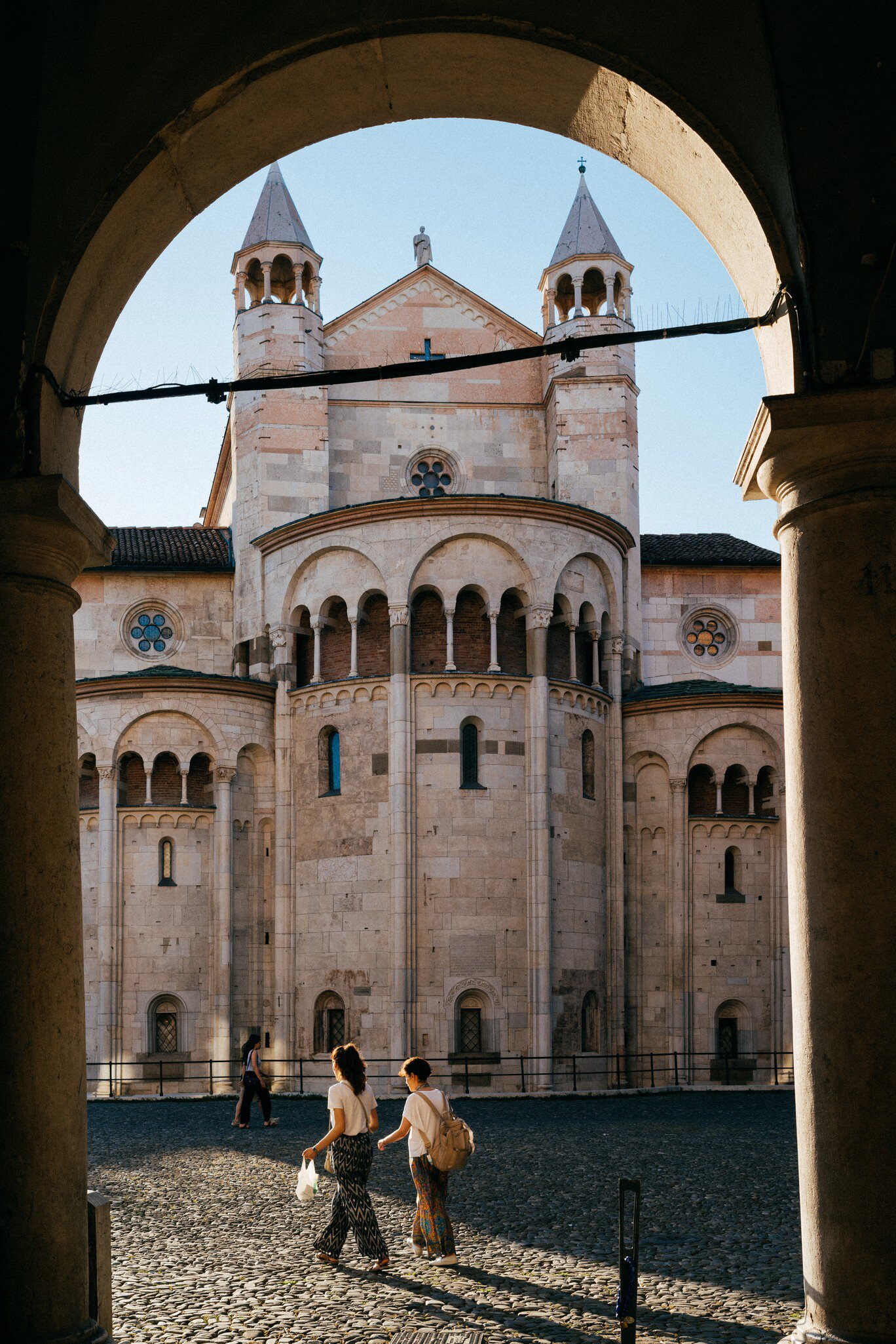 Two women cross a sunlit cobbled square towards the stone apse and twin spires of Modena's cathedral, seen through an arch.