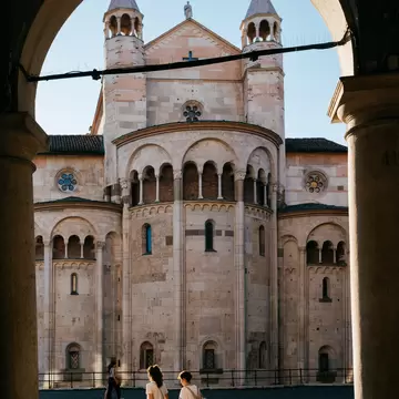 Two women cross a sunlit cobbled square towards the stone apse and twin spires of Modena's cathedral, seen through an arch.