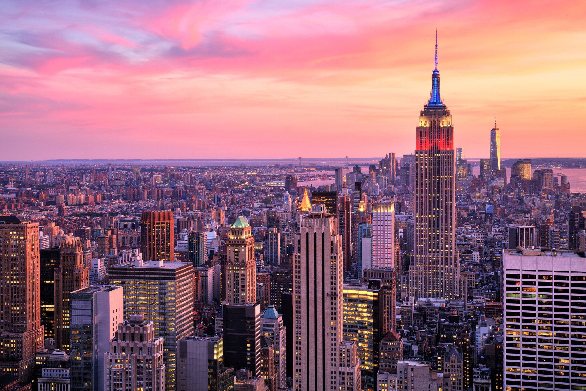 Floodlit tower crowned in red and blue rises above lit Midtown blocks, with water and pink clouds beyond.