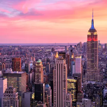 Floodlit tower crowned in red and blue rises above lit Midtown blocks, with water and pink clouds beyond.